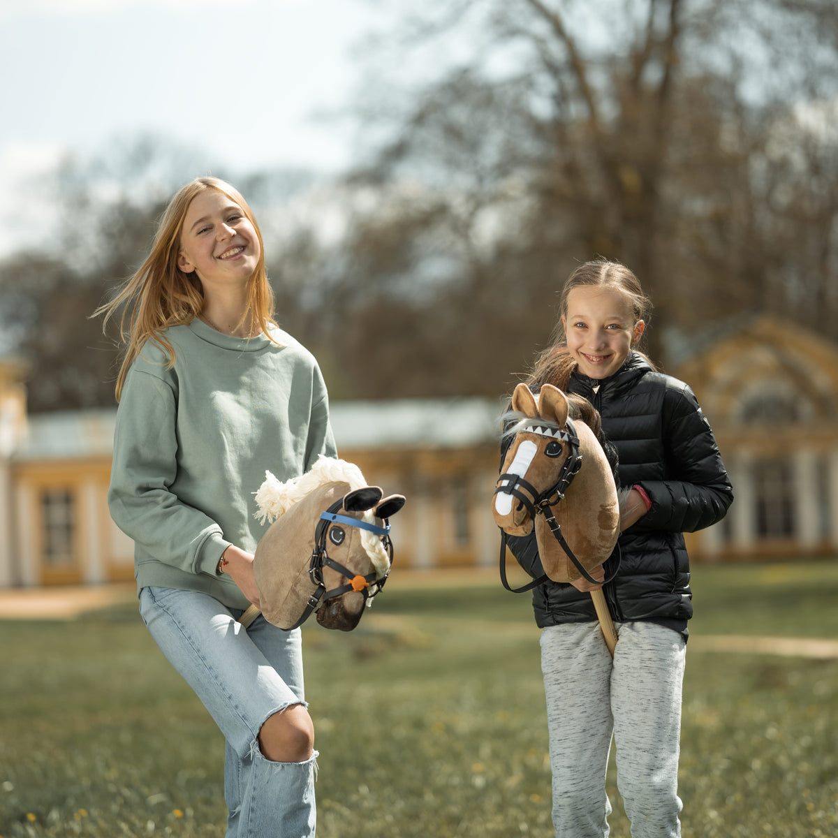 Deux filles souriantes dans un parc avec des Hobby Horses beige et brun clair aux brides noires, un bâtiment jaune derrière