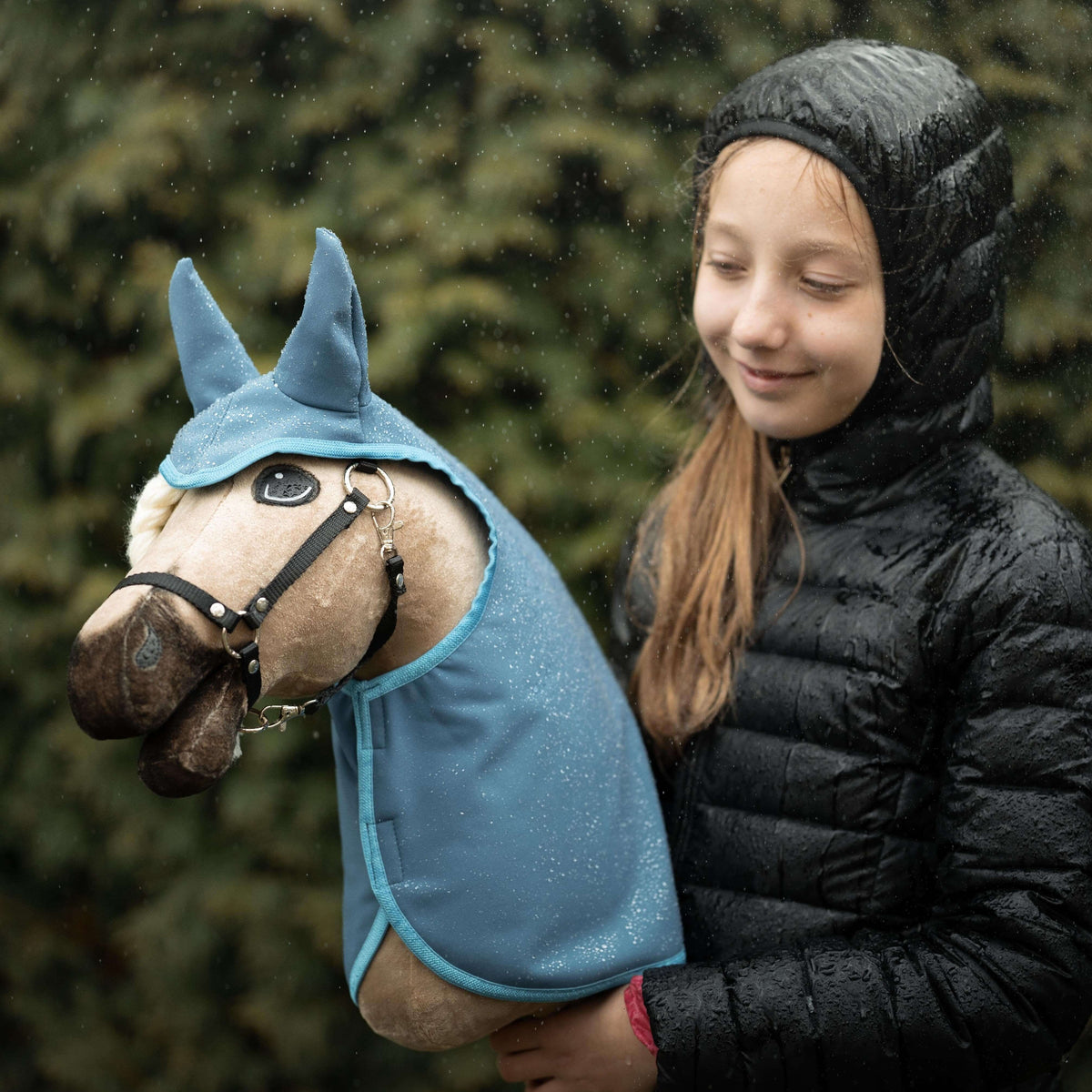 Fille souriante en doudoune noire sous la pluie avec un Hobby Horse beige vêtu d’un imperméable bleu pailleté à oreilles