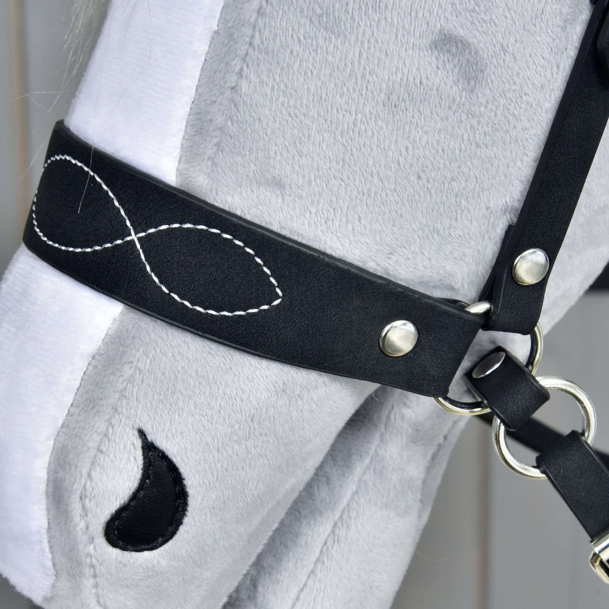 Close-up of the black noseband with white infinity-shaped stitching, silver rivets and rings on a grey plush muzzle