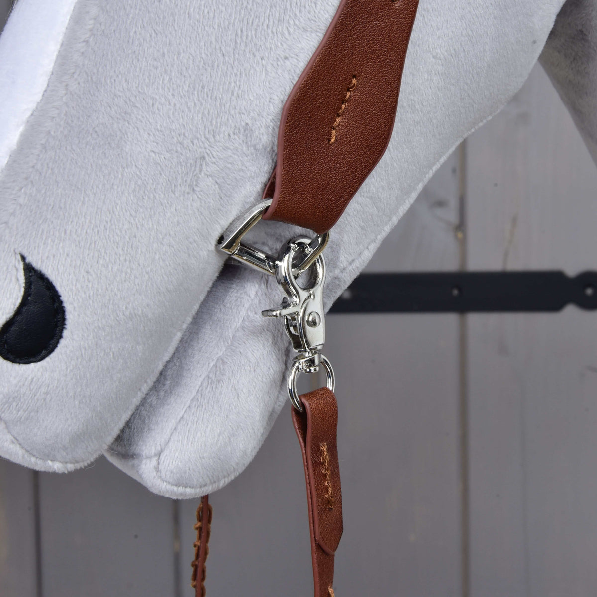Close-up of the silver snap hook clipping the brown rein to the cheekpiece ring at the muzzle of a grey plush head