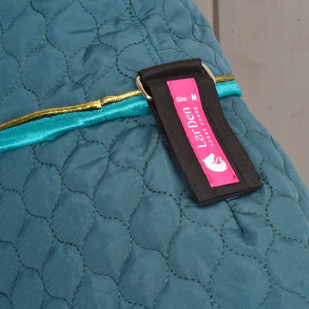 Close-up van een roze LarDen-label op een zwart riempje van de deken Lagoon, boven de turquoise rand en gouden bies