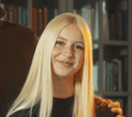 Smiling young woman with long blonde hair in a black top in front of a bookshelf