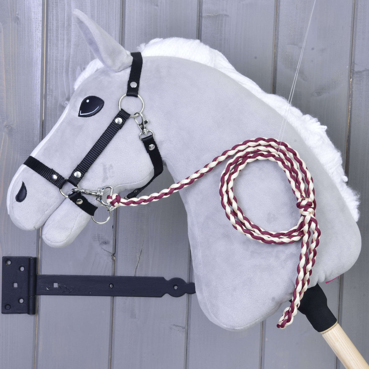 Maroon and cream braided lead rope clipped to the black halter of a grey Hobby Horse by a grey wooden wall