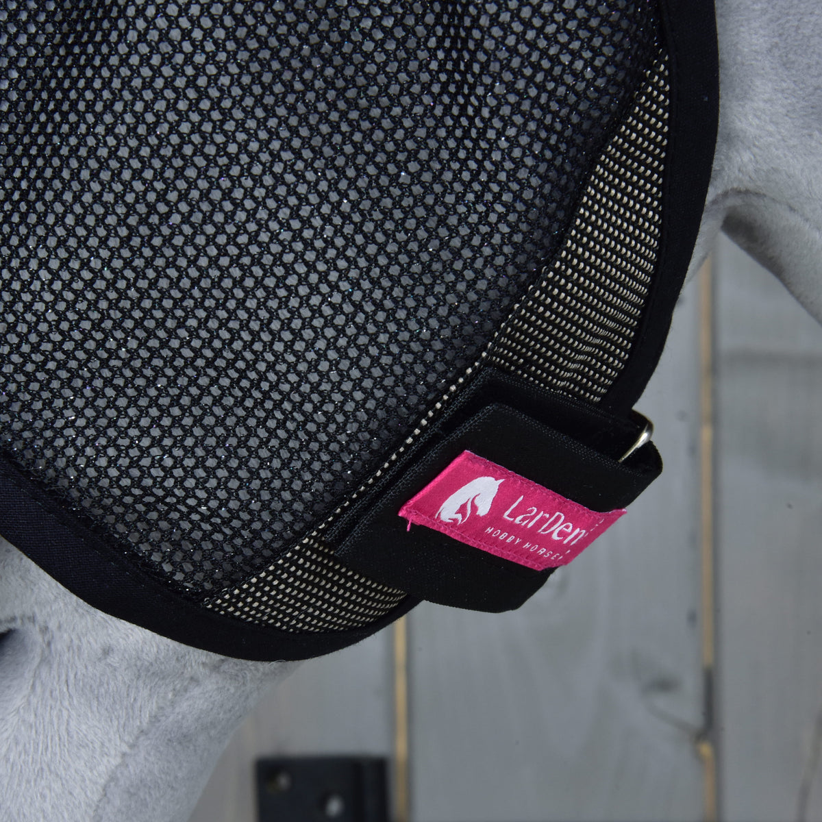 Close-up van het roze LarDen-label op de zwarte klittenbandsluiting bij de wang van het vliegenmasker op grijs pluche