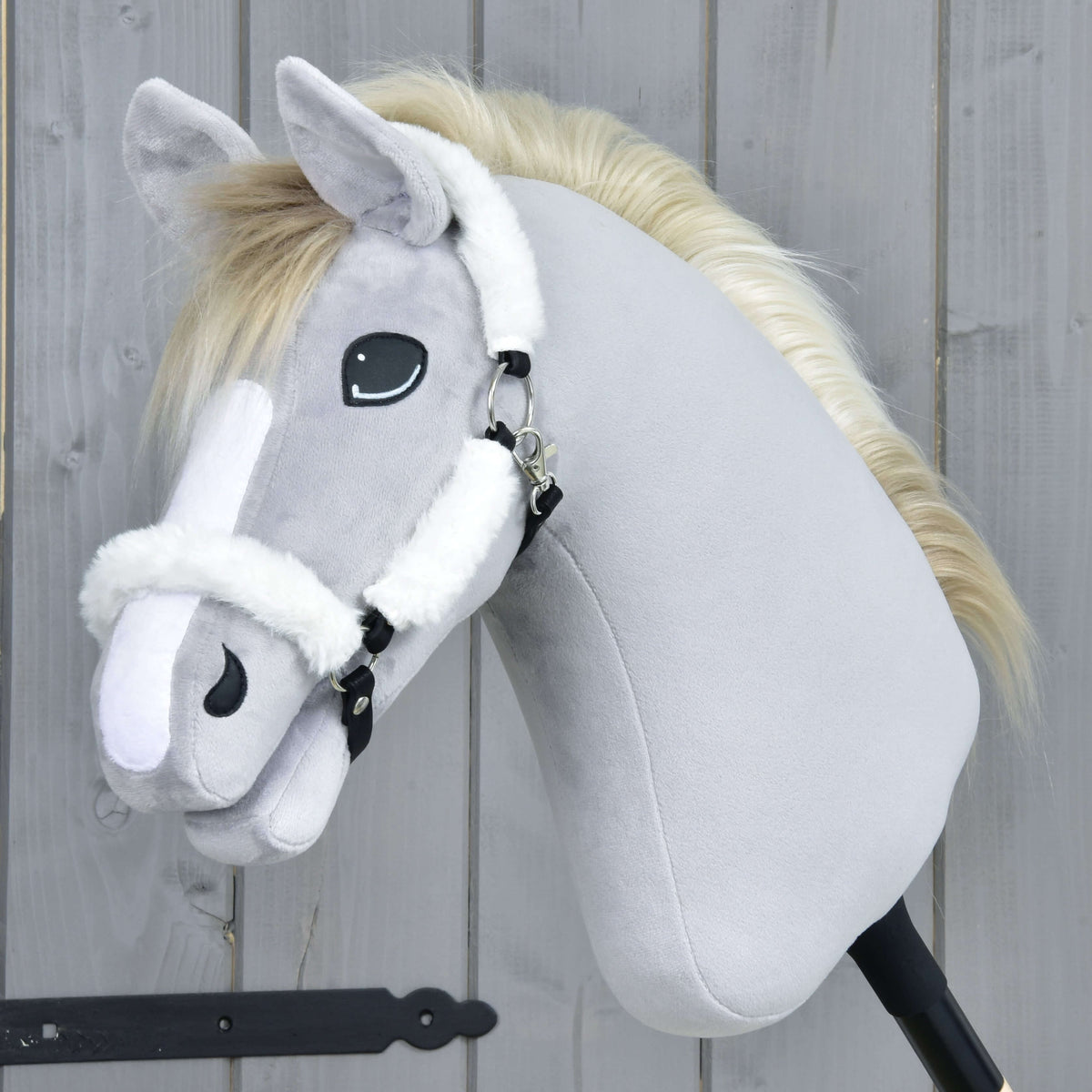 Grey plush Hobby Horse with a white blaze and cream mane in the white fur halter, side view by a grey wooden wall