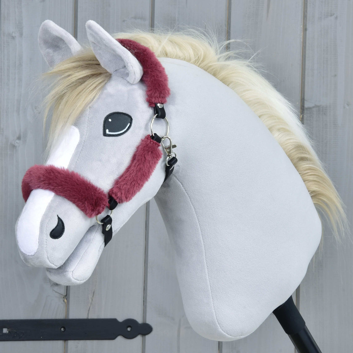 Grijze pluche Hobby Horse met crèmekleurige manen in de bordeauxrode halster met bont, zijaanzicht voor een grijze houten wand