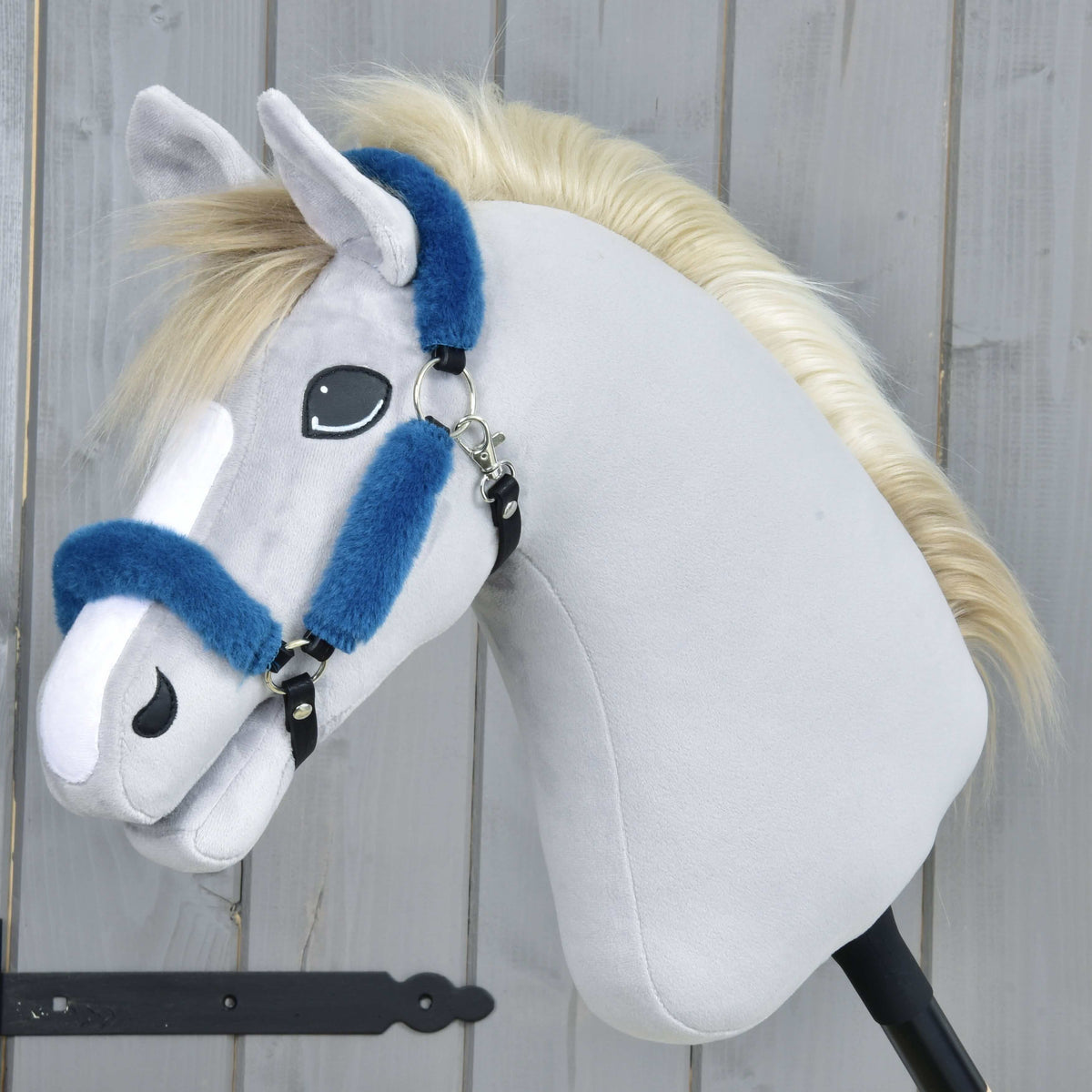 Grey plush Hobby Horse with a cream mane and white blaze wearing the petrol blue fur halter against a grey wooden wall