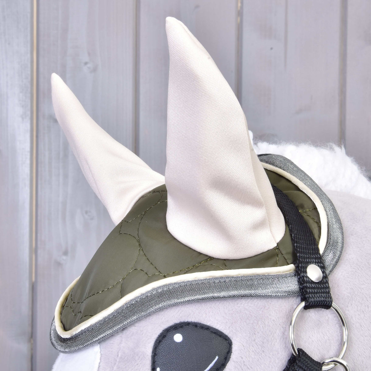 Gros plan sur le bonnet d’oreilles olive matelassé aux oreilles crème et passepoil gris de l’ensemble Olivetti, sur un Hobby Horse gris