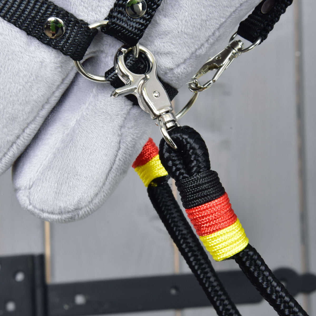 Close-up van de karabijnhaak van de Teugels Emblem aan de ring van de halster, met gele en rode omwikkeling op het zwarte touw