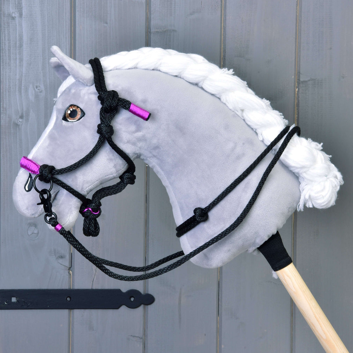 Un Hobby Horse gris en peluche à crinière blanche tressée porte le licol en corde noir et violet avec des rênes en corde noire