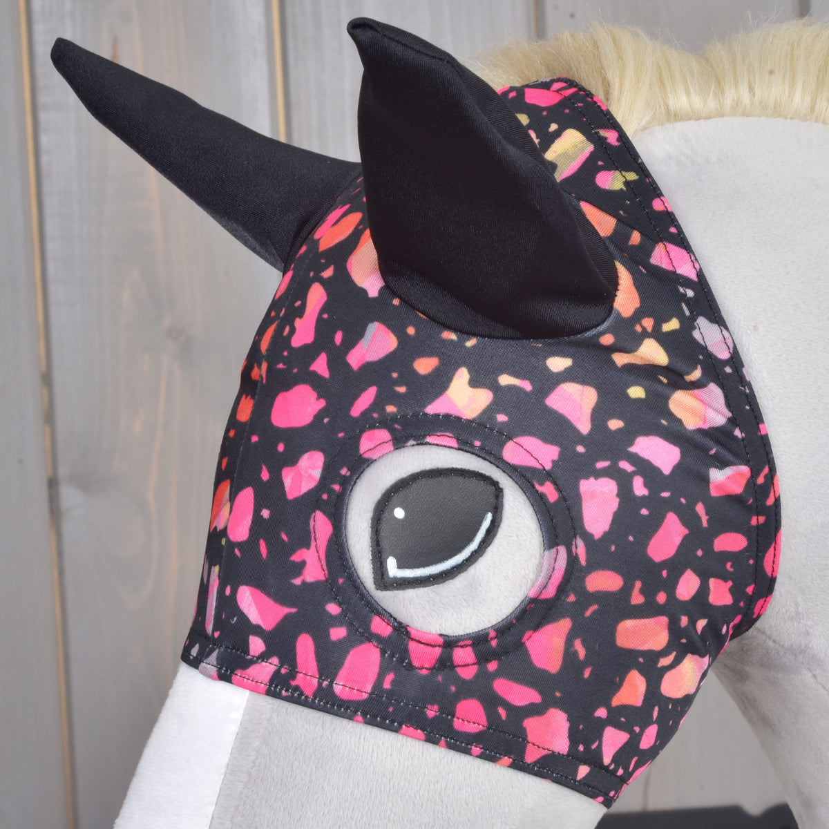 Gros plan sur le masque pour cheval Roxy porté par un Hobby Horse gris, oreilles noires et ouverture pour l’œil parmi les éclats rose et corail