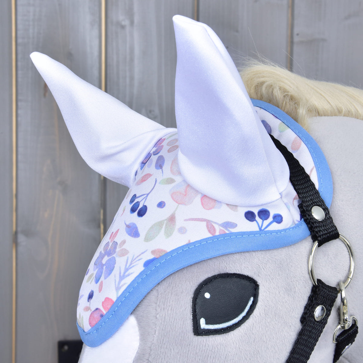 Close-up van het gebloemde oornetje met glanzende witte oren en blauwe rand op een grijze Hobby Horse naast het zwarte halster