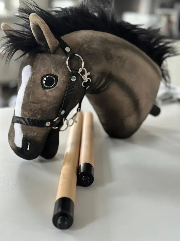 Tête de Hobby Horse marron à crinière noire, liste blanche et licol noir à côté d’un bâton en bois en deux parties aux embouts noirs
