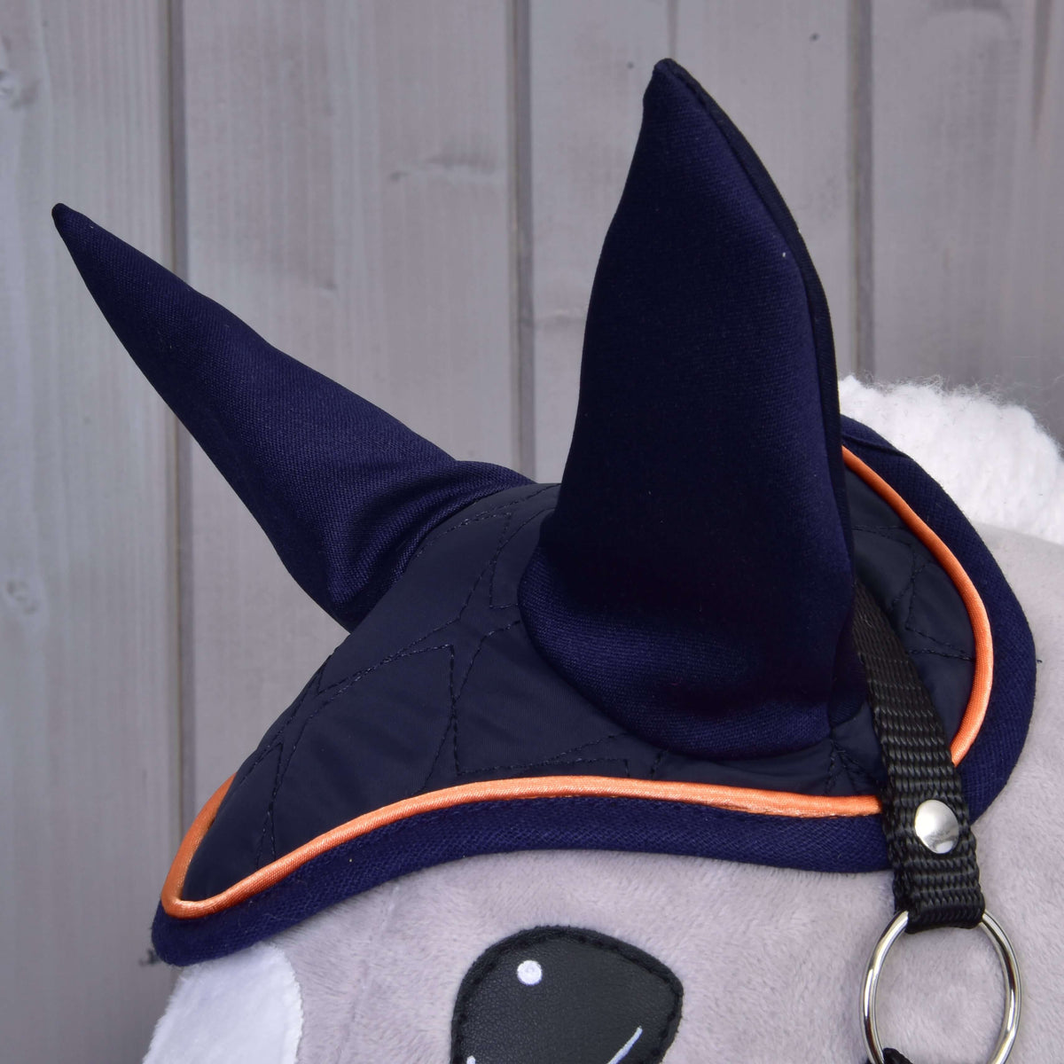 Gros plan sur le bonnet d’oreilles bleu marine aux hautes oreilles et passepoil orange sur un Hobby Horse gris