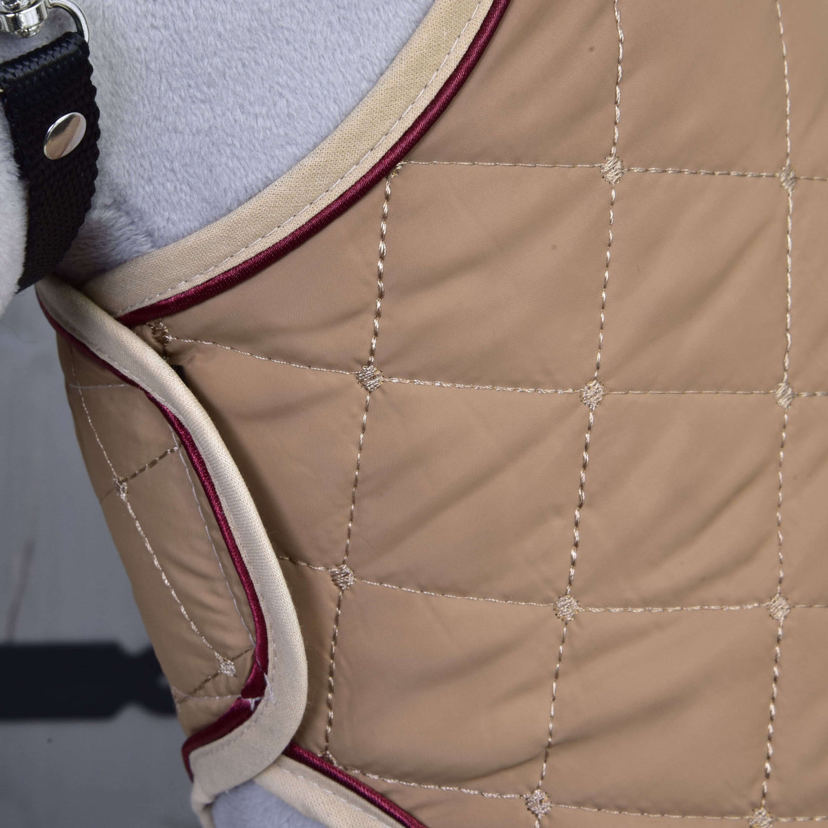 Gros plan sur la couverture beige matelassée en carrés, bordure crème et passepoil bordeaux sur un Hobby Horse gris
