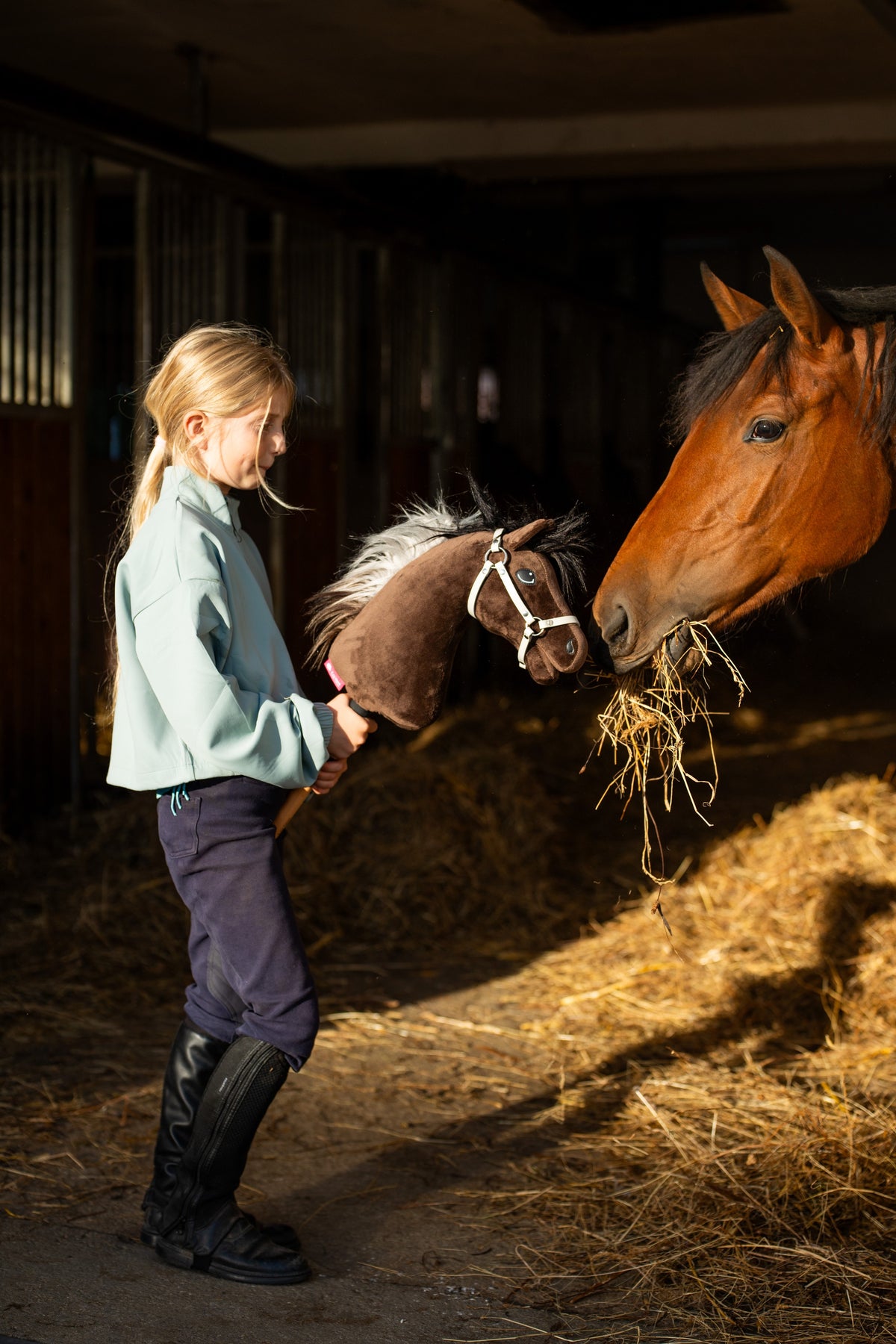 Fille en haut menthe et bottes d’équitation dans une écurie, tenant le Hobby Horse Alfredo au licol blanc près d’un cheval bai qui mange du foin