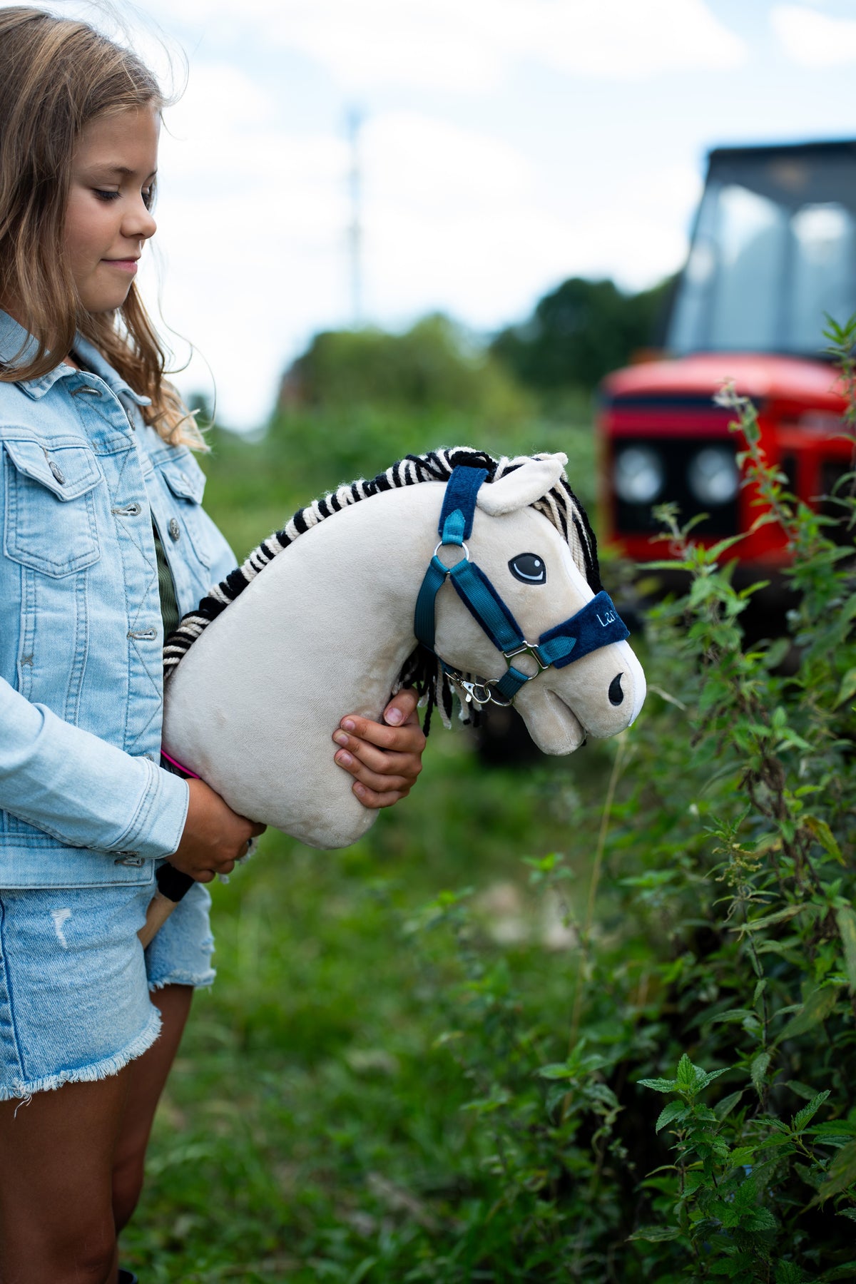 Fillette en veste en jean tenant Hobby Horse Aurora au licol bleu dehors près de plantes vertes, un tracteur rouge derrière