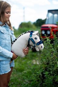Fillette en veste en jean tenant Hobby Horse Aurora au licol bleu dehors près de plantes vertes, un tracteur rouge derrière
