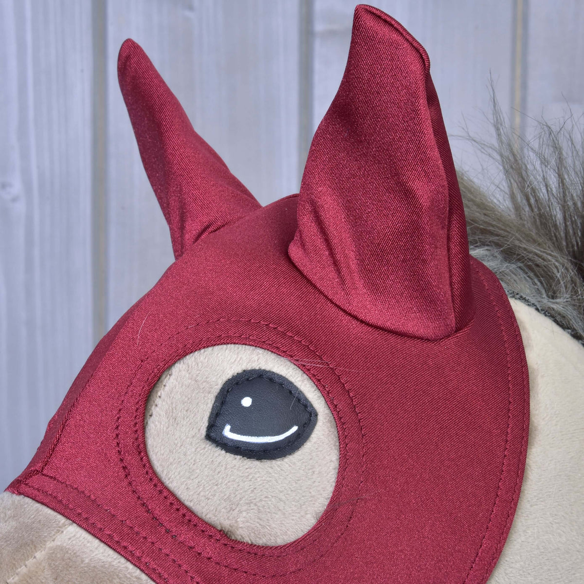 Detailopname van de glanzende donkerrode oorkapjes van het masker Barn Red met de gestikte oogopening eronder, op een beige Hobby Horse