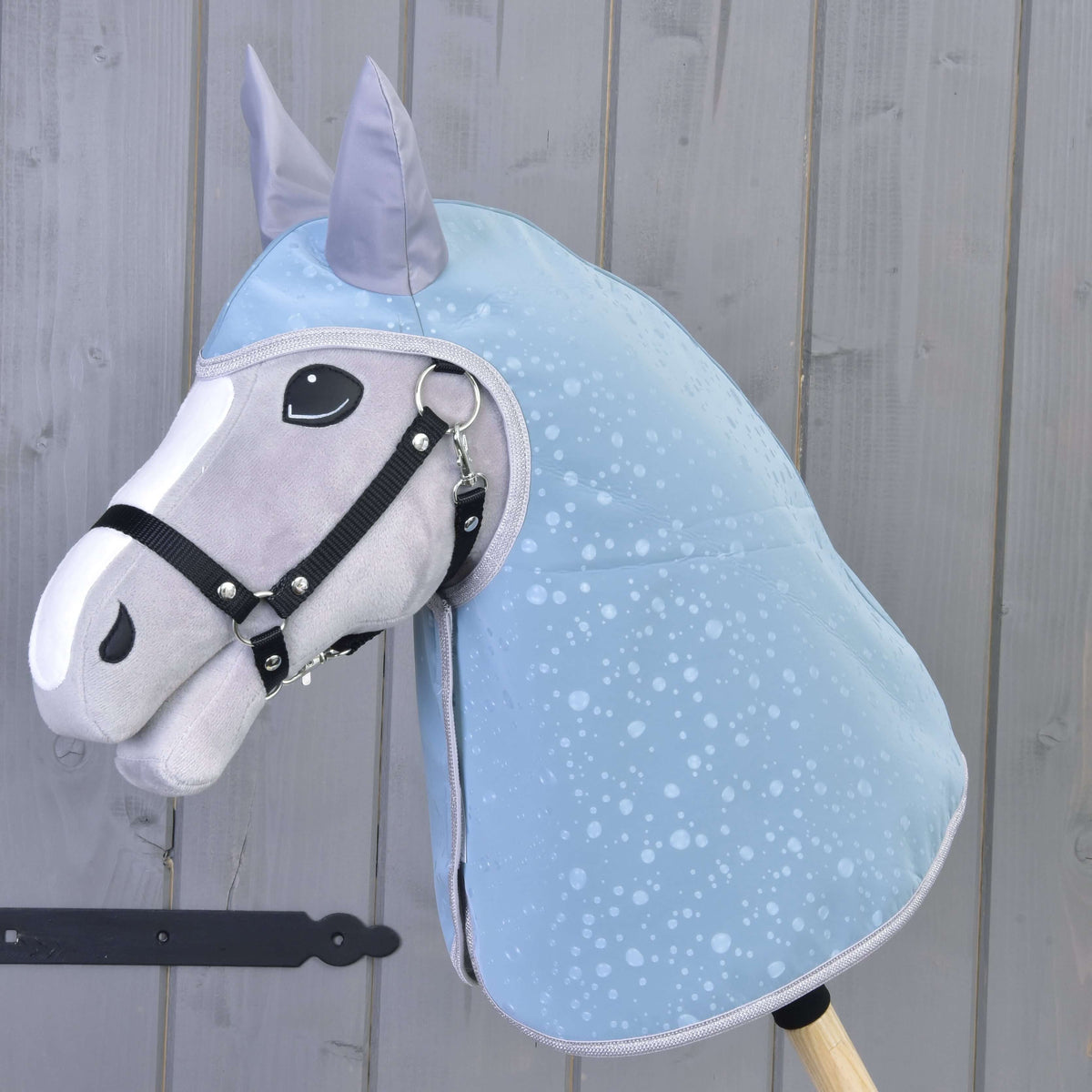 Regenjas Blue Drops op een grijze Hobby Horse met witte bles en zwart halster, bedekt hoofd en hals, voor een grijze houten wand