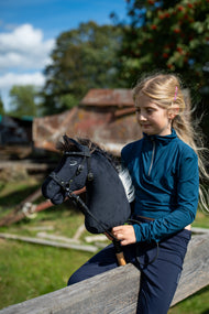 Fille blonde en haut bleu canard assise sur une barrière en bois avec le Hobby Horse Bond avec licol noir, ici avec bride noire et rênes