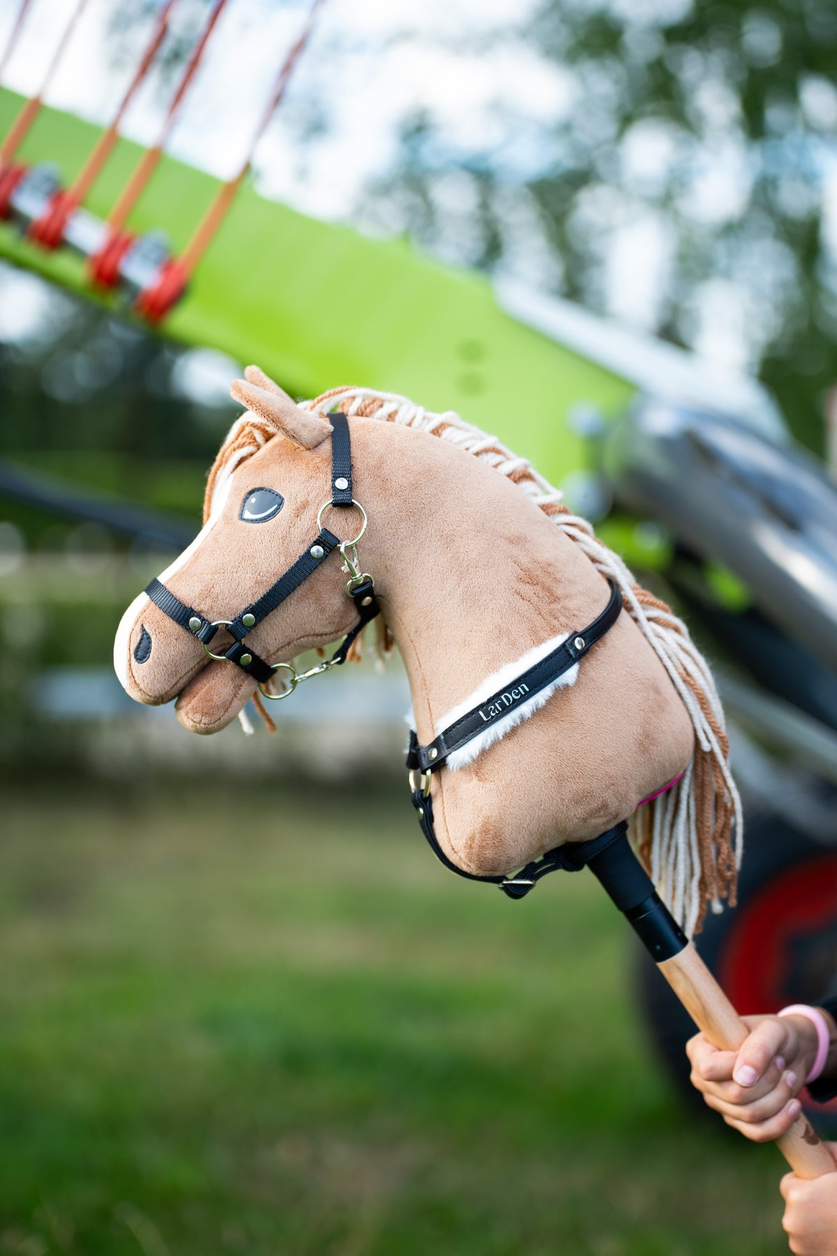 Hobby Horse Bren in a black halter and a black breastgirth with white fur and LarDen lettering, green farm machine behind