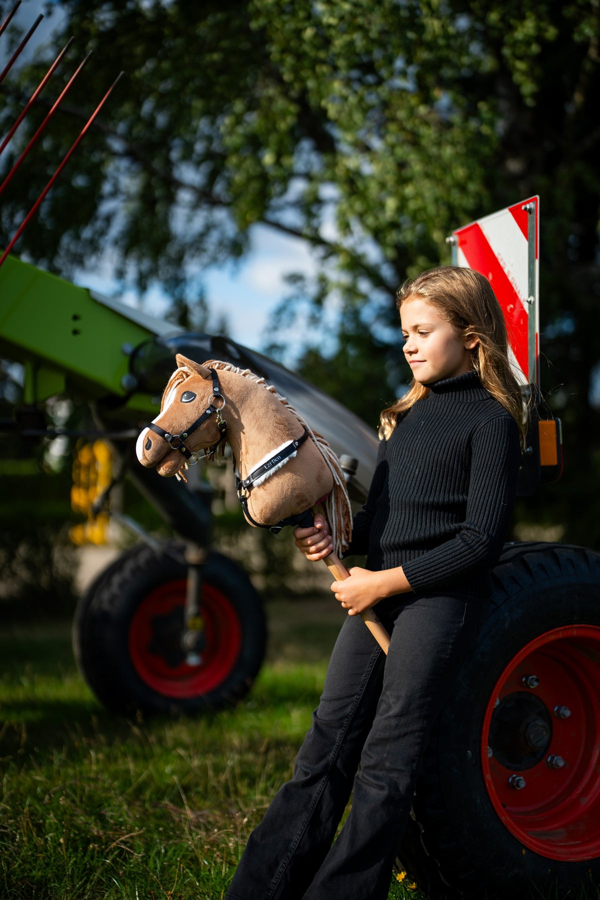 Girl in a black turtleneck by green farm machinery holds Hobby Horse Bren with Black Halter, here with a black bridle