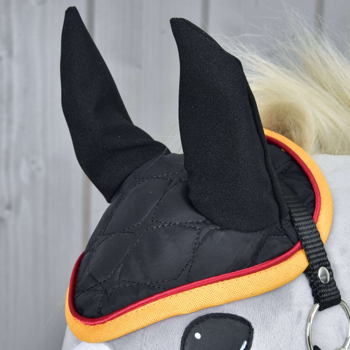 Gros plan du bonnet d’oreilles Flag DE sur un Hobby Horse gris : oreilles et matelassage noirs bordés de rouge et de jaune or
