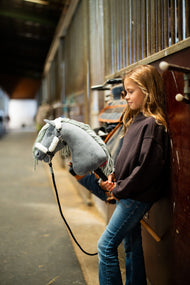Girl in a dark sweatshirt in a stable aisle holds a grey Hobby Horse in the white fur halter and a black lead rope