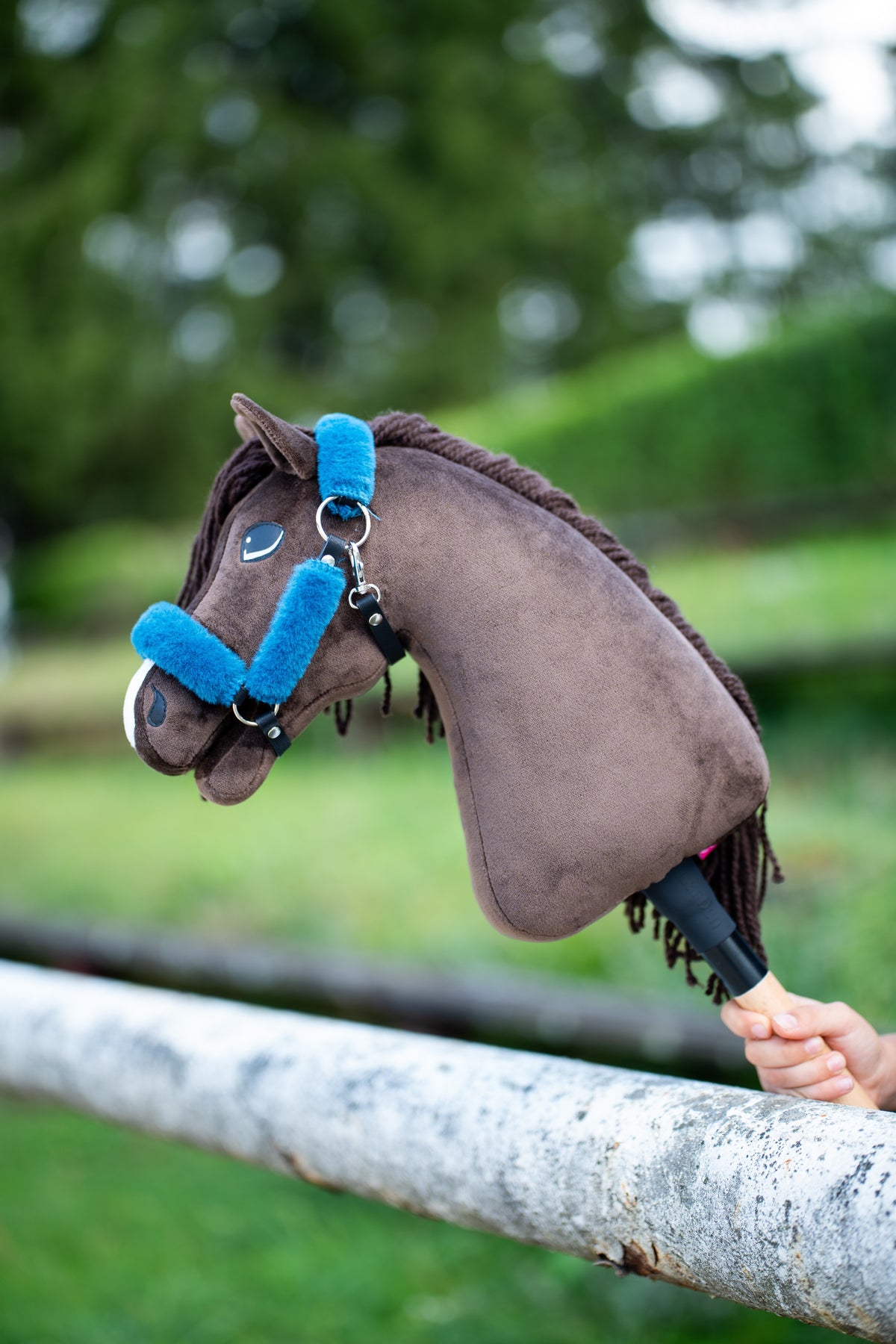 Hobby Horse Chianti met zwart halster met petrolblauw bont, rustend op een witte berkenstam voor groene struiken