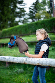 Meisje in een donkerblauwe bodywarmer bij een wit hek met de donkerbruine Hobby Horse in een blauw halster met bont, haag erachter