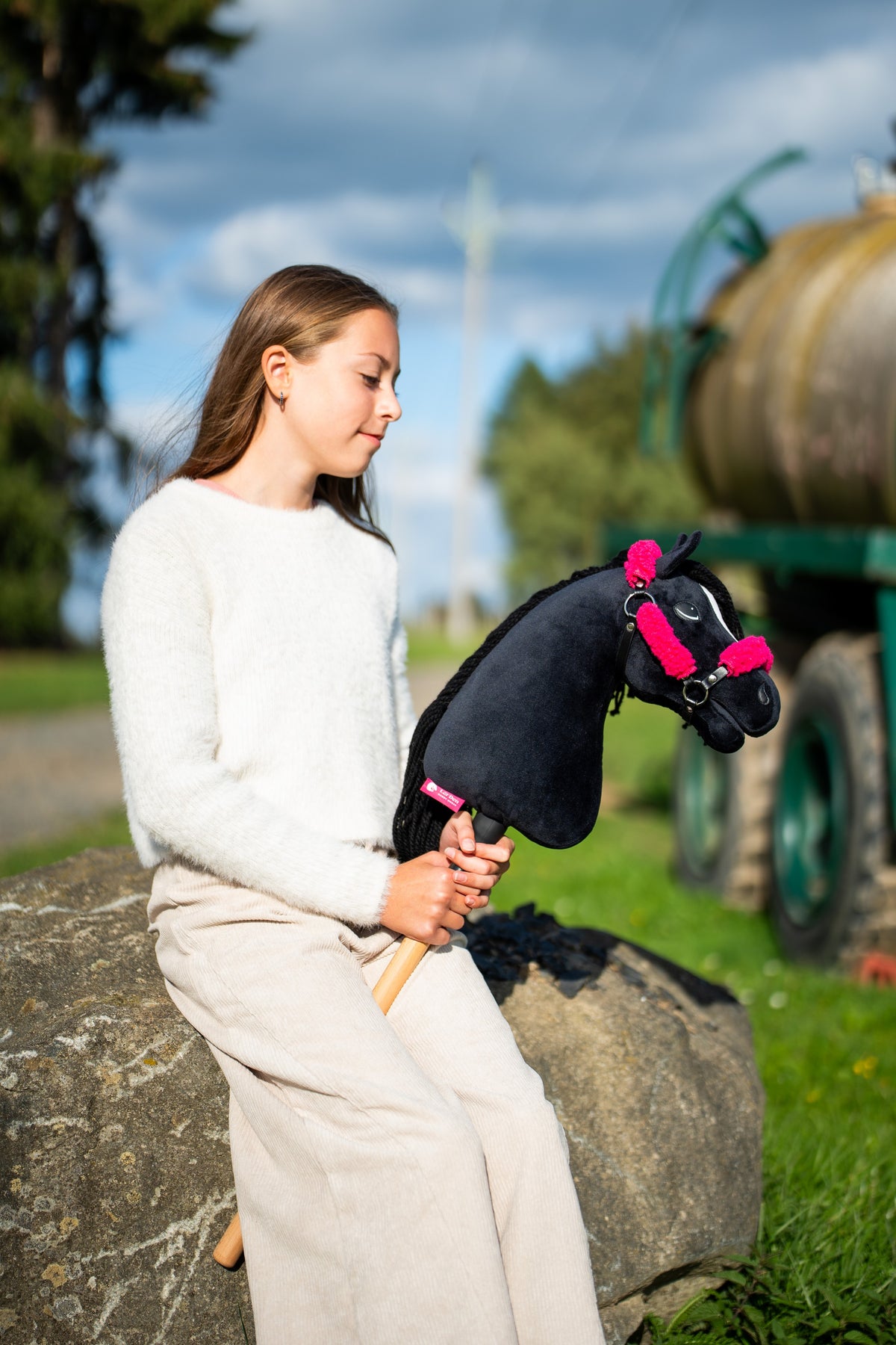 Girl in a cream jumper sitting on a large rock outdoors with a black Hobby Horse in the black halter with pink fur