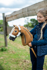 Fille en gilet bleu marine près d’une clôture en bois, tenant le Hobby Horse fauve doré au licol à fourrure blanche