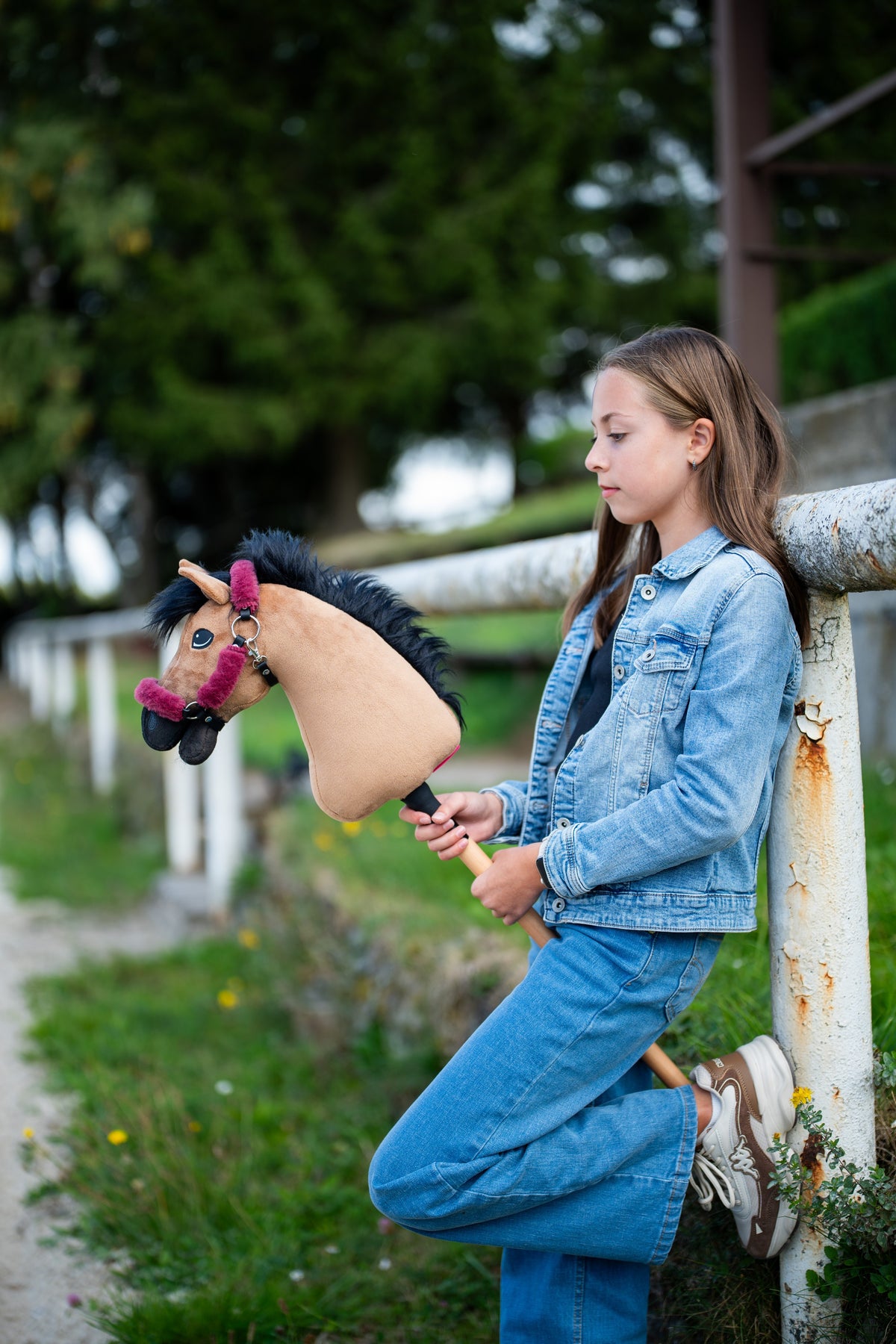 Meisje in spijkerjasje leunt tegen een wit hek en houdt de lichtbruine Hobby Horse vast in een frambozenrood halster met bont