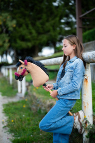 Meisje in spijkerjasje leunt tegen een wit hek en houdt de lichtbruine Hobby Horse vast in een frambozenrood halster met bont