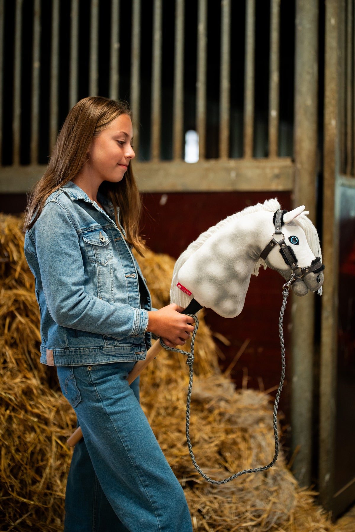 Meisje in spijkerjasje bij strobalen houdt Hobby Horse Elektra vast, hier met donkergrijze halster met bont en gevlochten grijs halstertouw