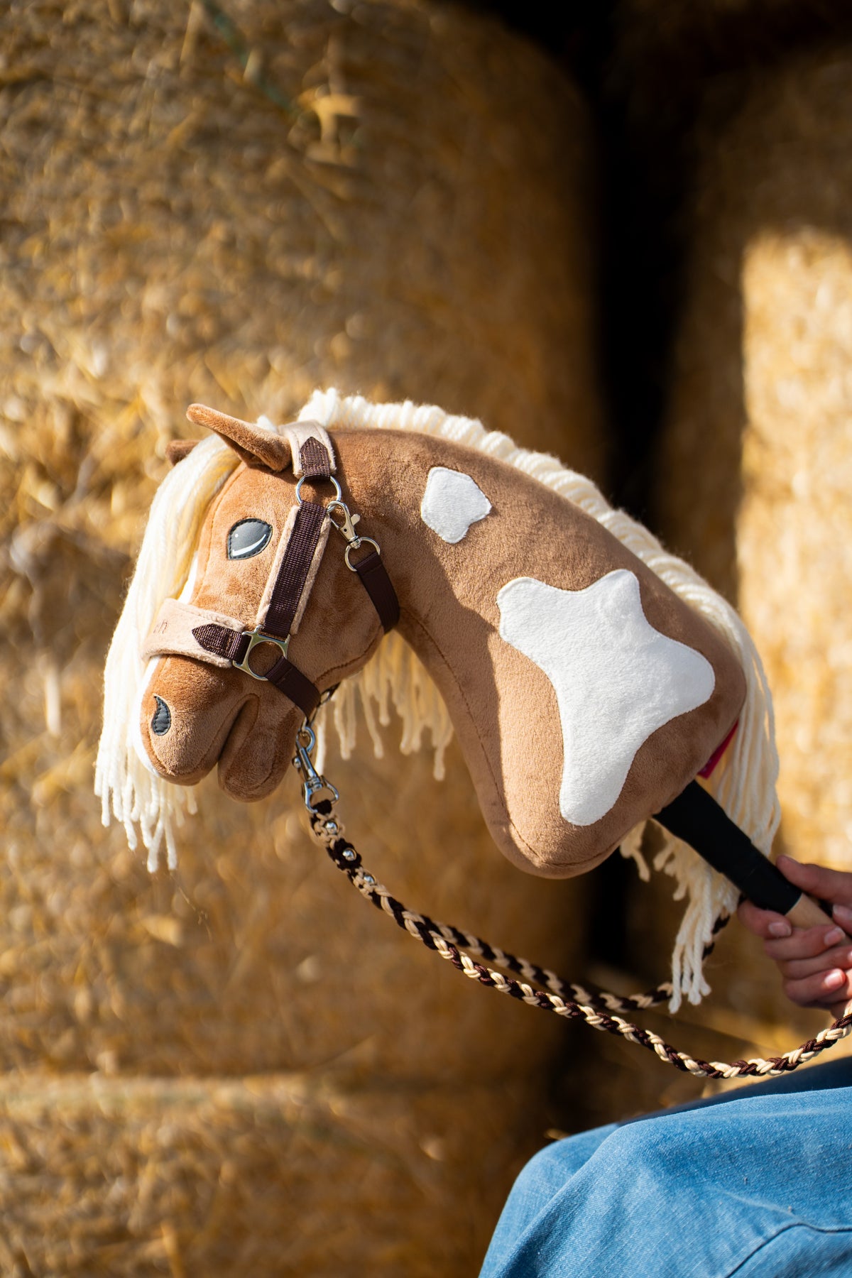 Hobby Horse Fury with white patches in a brown halter and a brown and cream braided lead rope, straw bales behind