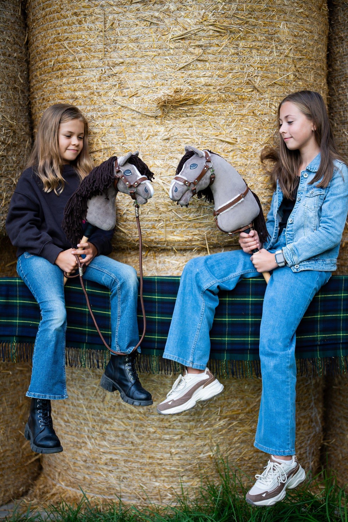 Twee meisjes op strobalen met een groen geruit plaid, elk met een grijze Hobby Horse in een bruine halster