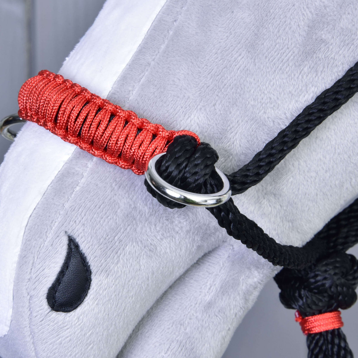 Close-up of the red braided noseband and silver ring of the black rope halter on a grey plush Hobby Horse