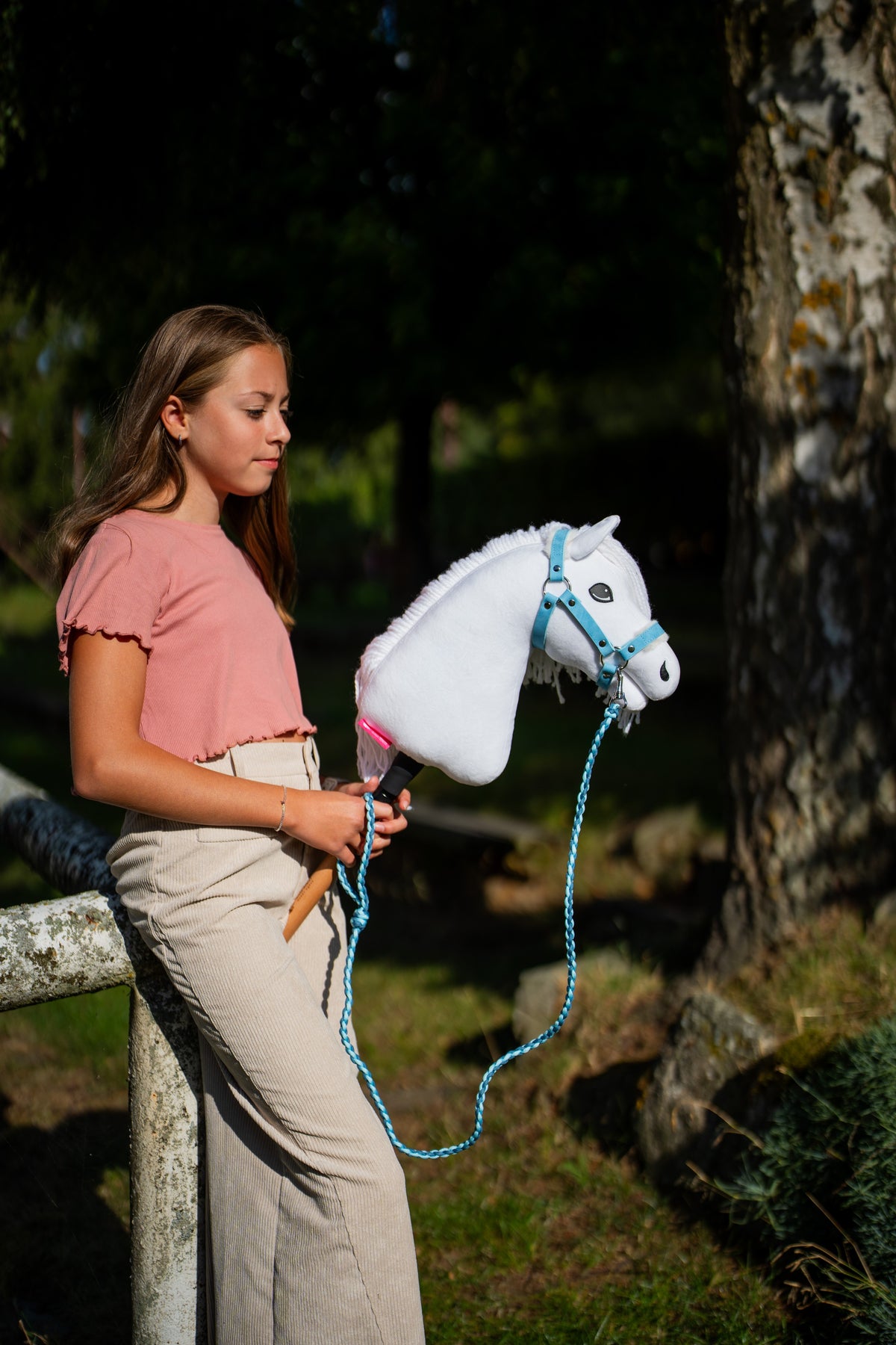 Girl in a pink top by a birch tree holding a white Hobby Horse in a pastel blue halter, the lead rope hanging in a loop