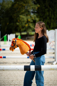 Meisje in zwart topje in een rijbak met hindernissen, met een vosbruine Hobby Horse in een zwart halster met wit bont