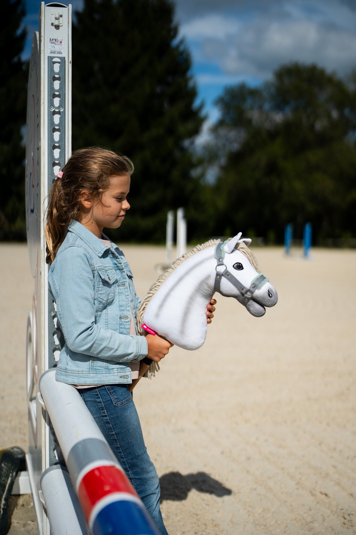 Een meisje in een spijkerjasje leunt tegen een hindernisstandaard in de rijbak en houdt een witte Hobby Horse met de grijze halster met mint bont vast