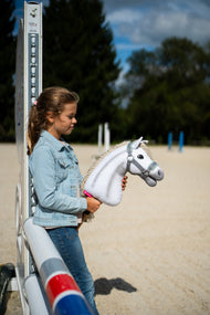 Une fille en veste en jean s’appuie sur un chandelier d’obstacle en carrière et tient un Hobby Horse blanc au licol gris à fourrure menthe