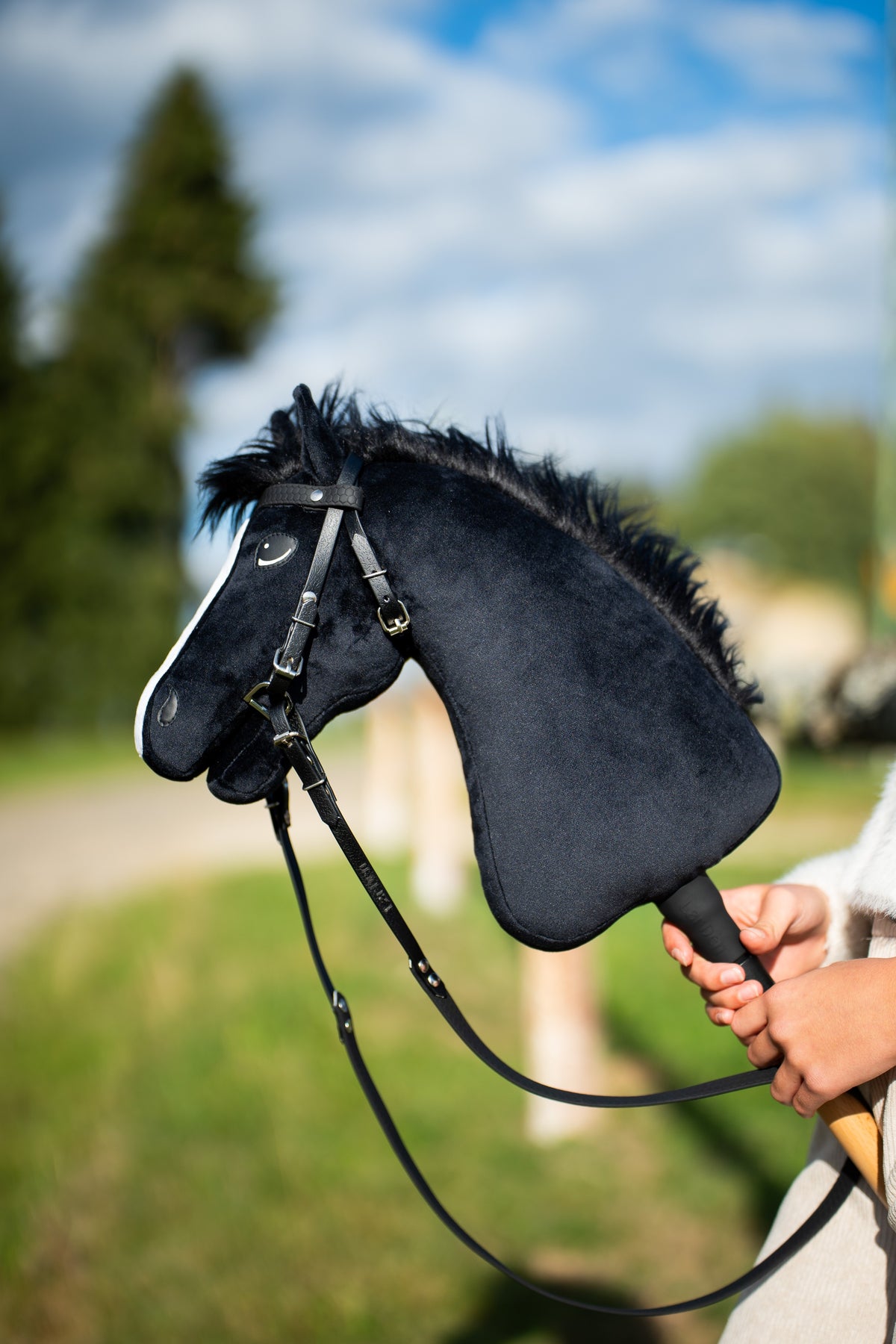 Hobby Horse Loki à crinière noire hirsute avec bride noire en similicuir et longues rênes, ciel bleu et sapin derrière