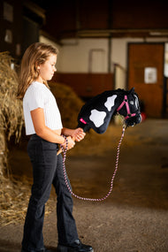 Meisje in wit shirt staat in een stal met Hobby Horse Merlin met zwart halster, hier met roze halster en gevlochten halstertouw