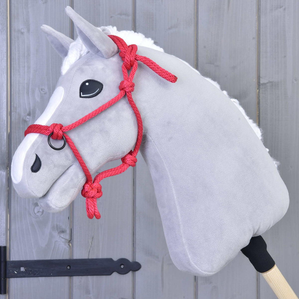 Grijze pluche Hobby Horse met witte bles en manen in het rode touwhalster voor een grijze houten wand