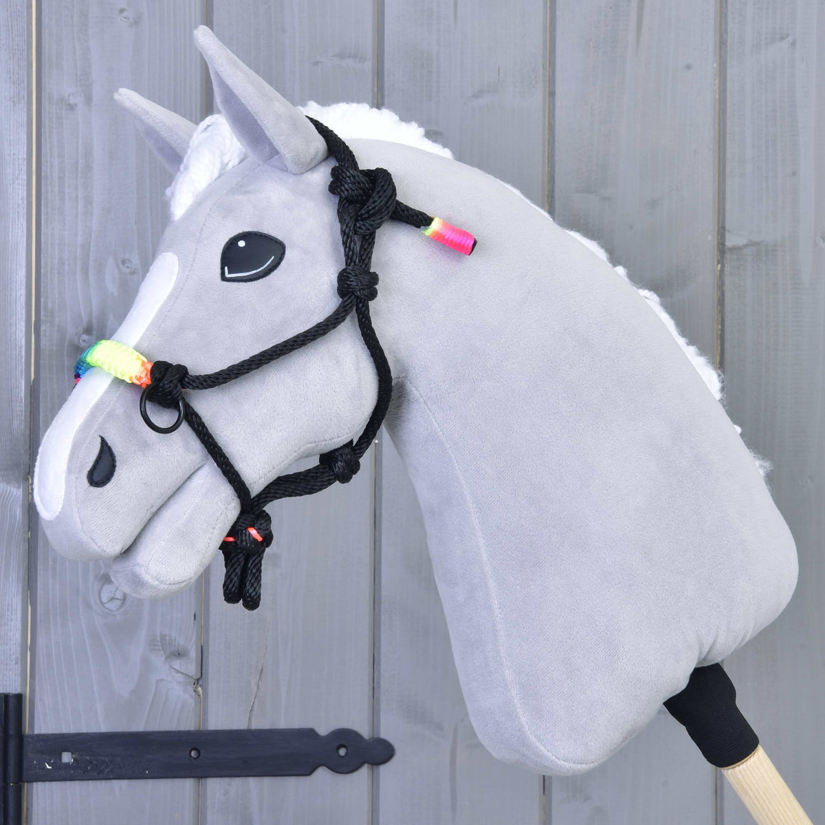 Grey plush Hobby Horse with a white mane wearing the black rope halter with rainbow noseband in front of a grey wooden wall