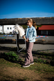 Jong meisje in spijkerjasje bij een houten hek houdt Hobby Horse Pixie vast in een grijze halster met bont en halstertouw