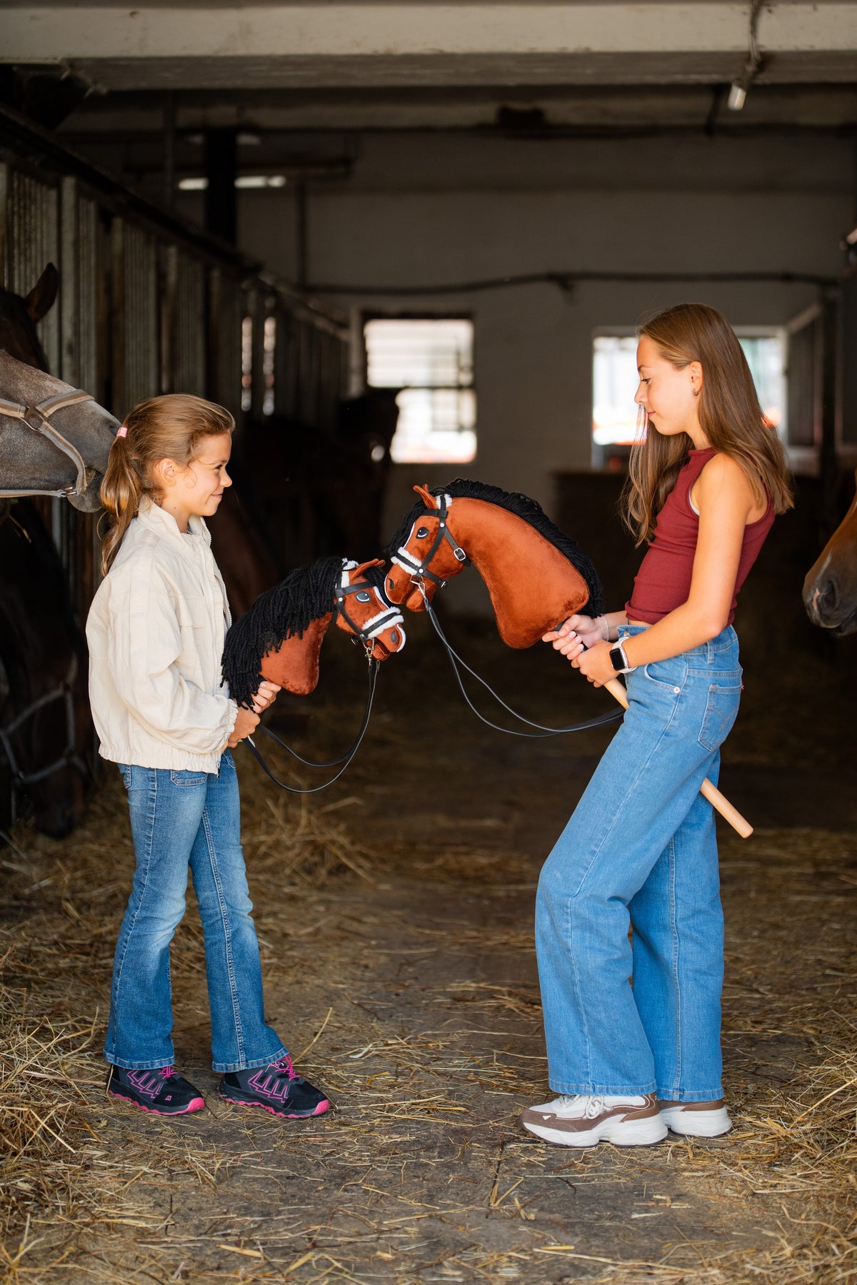Two girls in a stable aisle, each holding a black-maned chestnut Hobby Horse in a black bridle with white padding