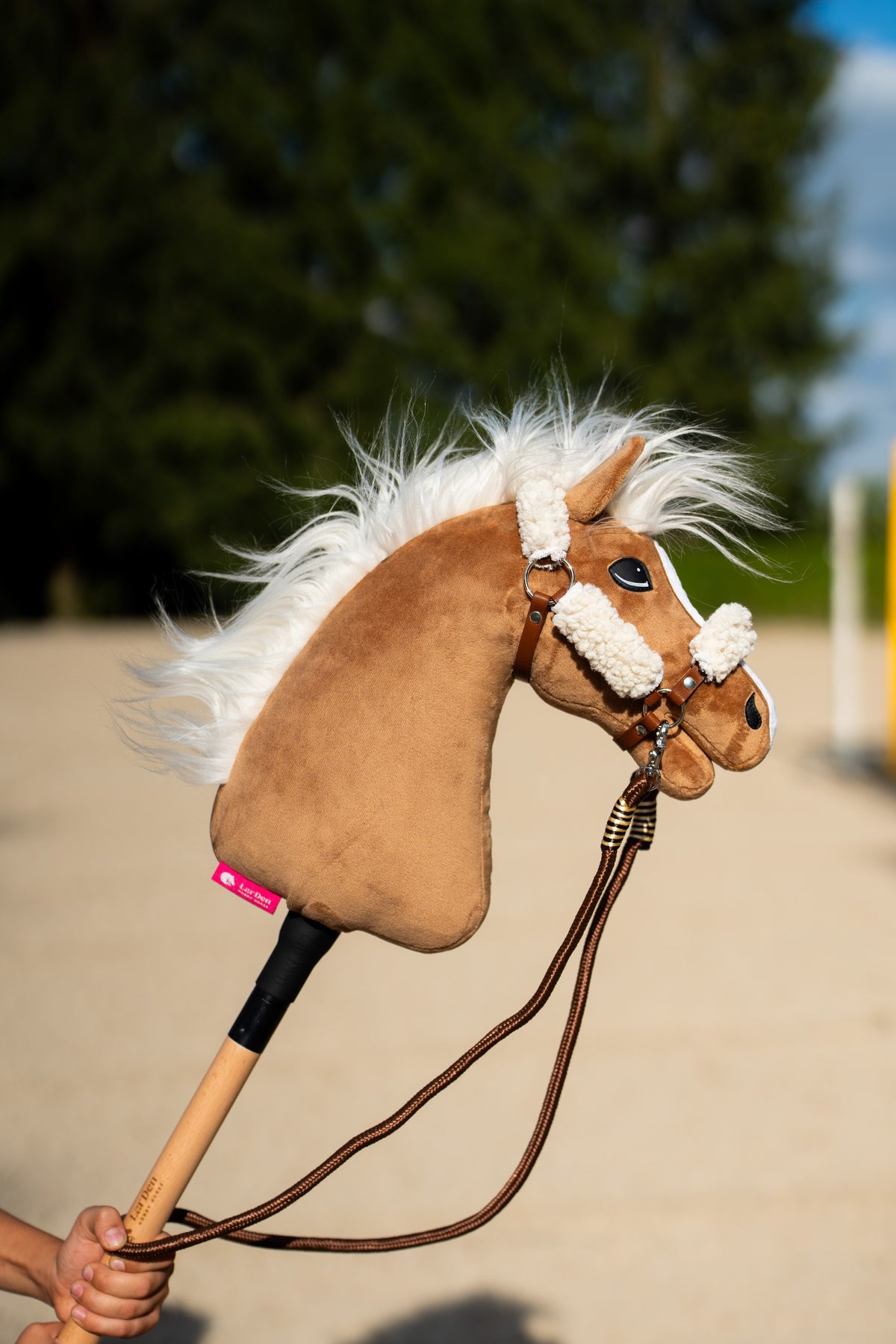 Hobby Horse Star met bruine halster met crèmekleurig bont en bruine touwteugels in een kinderhand op een zonnige rijbaan